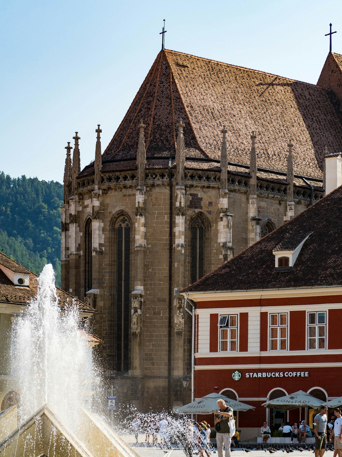The Black Church rising above Piața Sfatului in Brașov, a fountain throwing spray in the foreground and a forested hillside behind