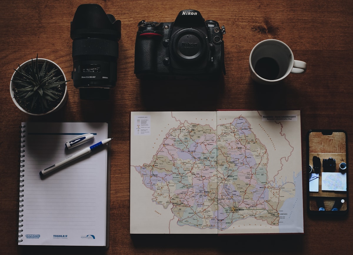 A road map of Romania open on a wooden table beside a camera, a cup of coffee, a notebook and a phone